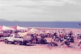 Faded photograph of three tents, a truck and lots of motorbikes on the beach on a sunny day. 