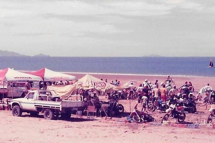 Faded photograph of three tents, a truck and lots of motorbikes on the beach on a sunny day. 