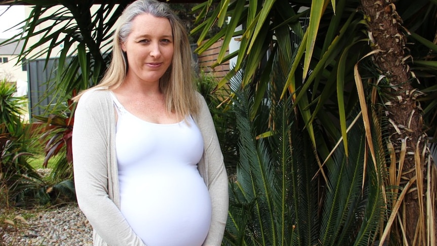 A heavily pregnant woman stands among a leafy yard.