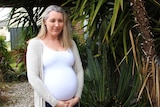 A heavily pregnant woman stands among a leafy yard.