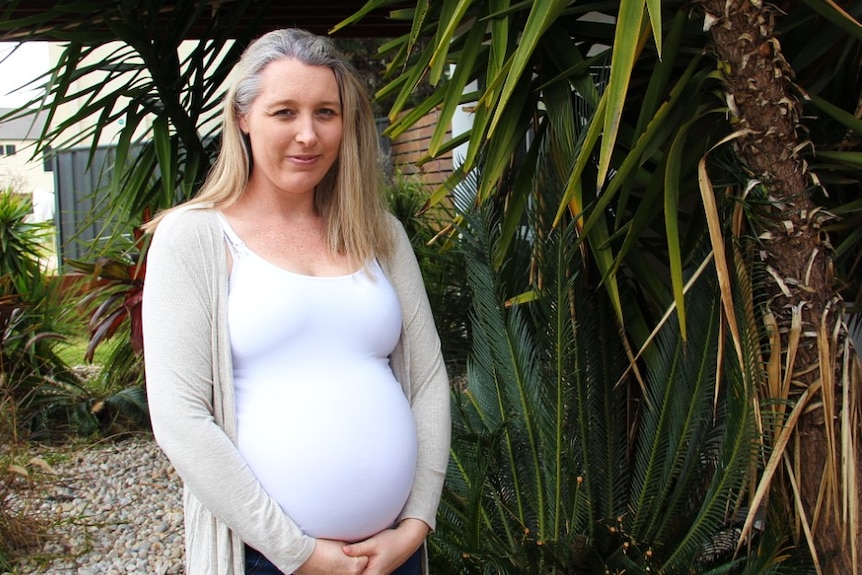 A heavily pregnant woman stands among a leafy yard.