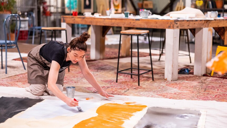 Bronwyn knees on the ground, leaning over a large canvas on which she is painting large blobs of orange and white