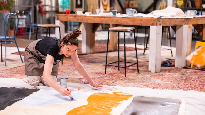 Bronwyn knees on the ground, leaning over a large canvas on which she is painting large blobs of orange and white