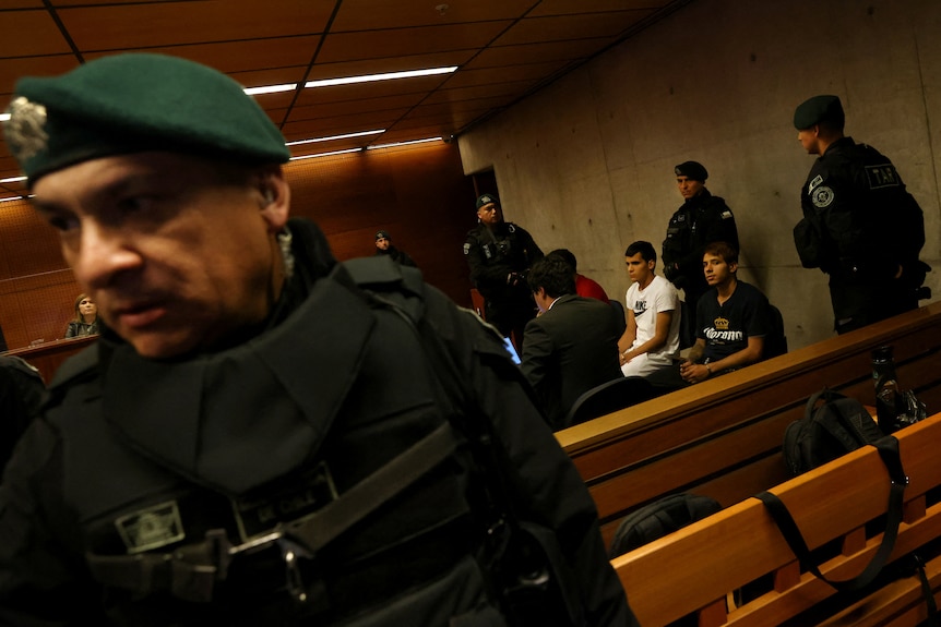 A group of young men sit handcuffed in a courtroom surrounded by soldiers in green berets.