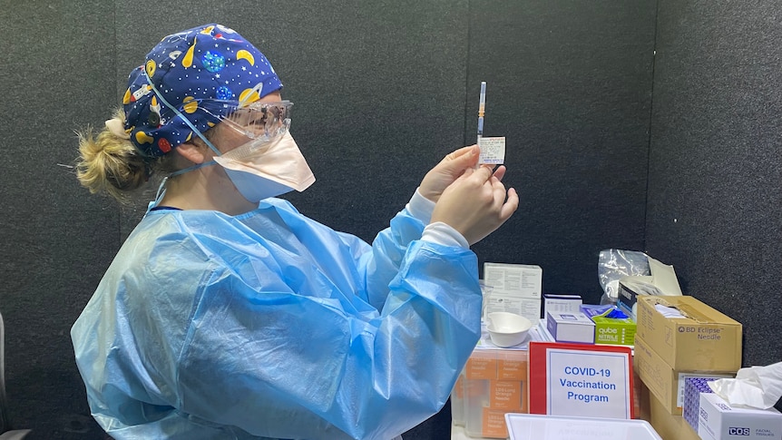 A woman in blue PPE holds up a syringe containing Pfizer vaccine.