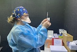 A woman in blue PPE holds up a syringe containing Pfizer vaccine.