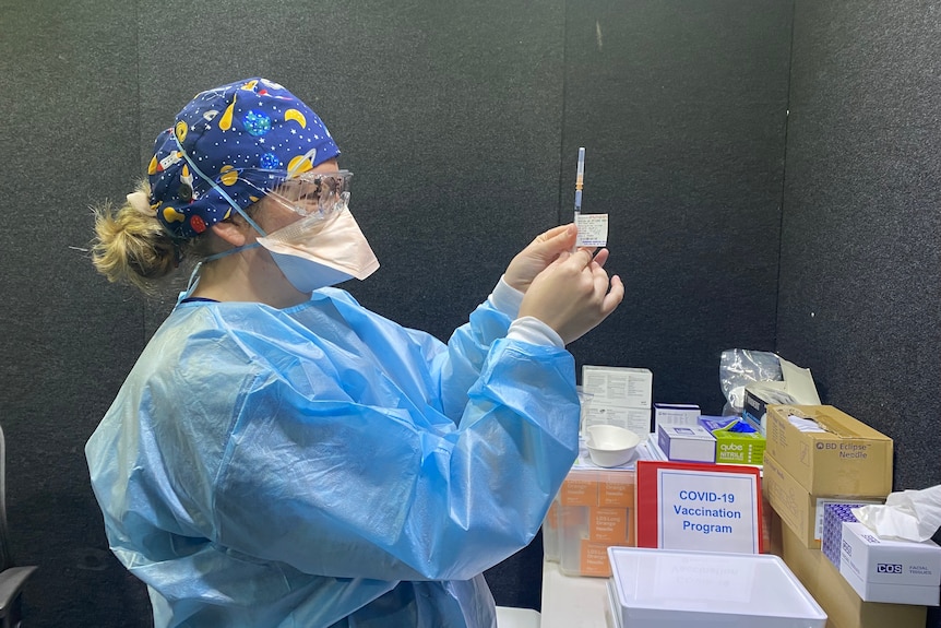 A woman in blue PPE holds up a syringe containing Pfizer vaccine.