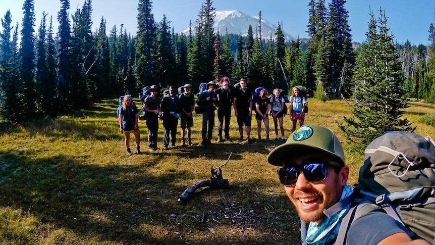 A smiling young man with a trail backpack smiles as he poses for a photo with a group of students lined up in a forest clearing.