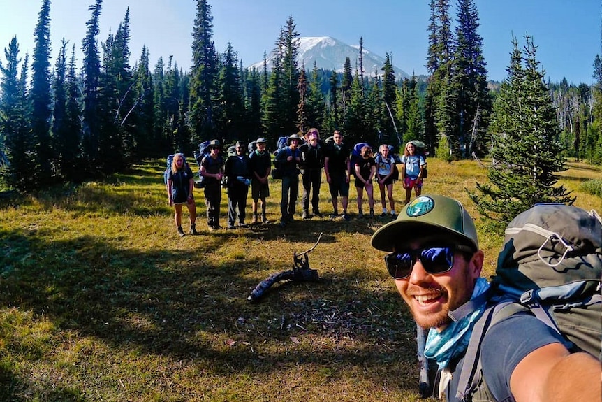 A smiling young man with a trail backpack smiles as he poses for a photo with a group of students lined up in a forest clearing.