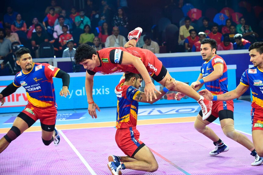 A kabaddi player wearing red leaps over a competitor as other players and an arena crowd watch on.