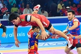A kabaddi player wearing red leaps over a competitor as other players and an arena crowd watch on.