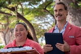 A short smiling Hawaiian woman holding a tray of white towels stands next to a tall smiling white man holding a black folder