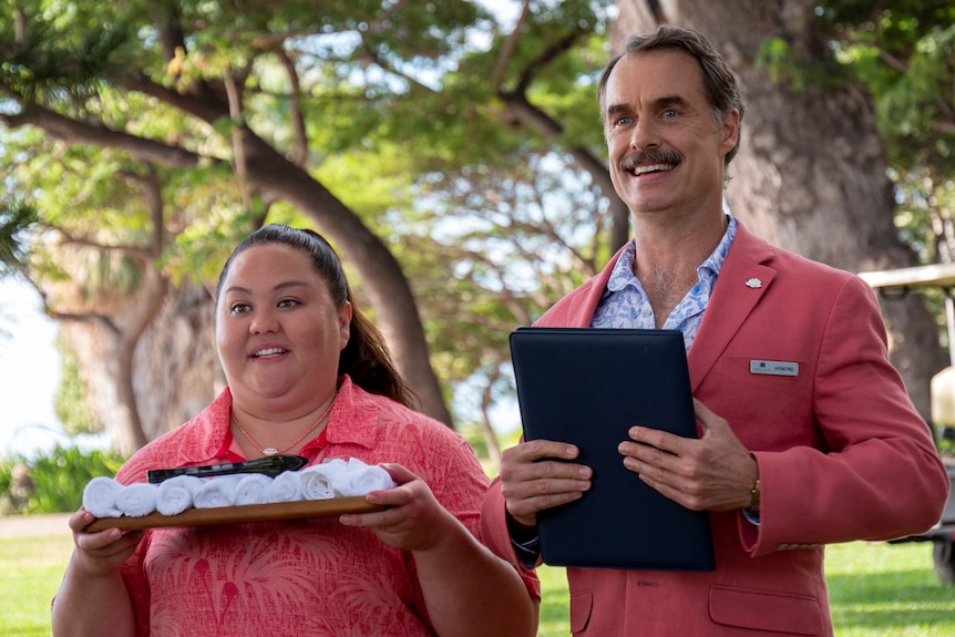 A short smiling Hawaiian woman holding a tray of white towels stands next to a tall smiling white man holding a black folder
