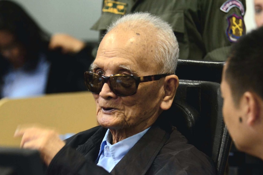 Nuon Chea, an old man, sits in a courtroom wearing dark glasses. There is a guard standing behind him.
