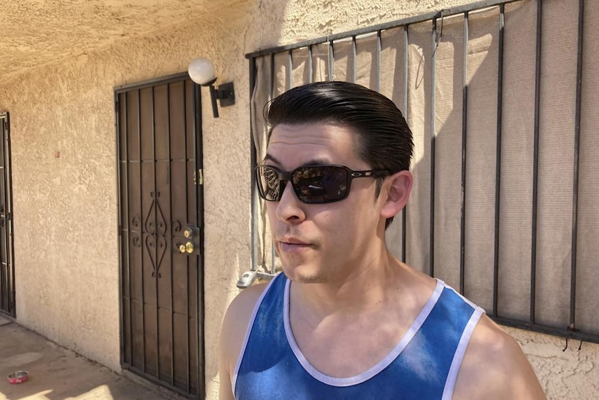 Hispanic man with slicked back black hair, wearing a blue singlet and black sunglasses