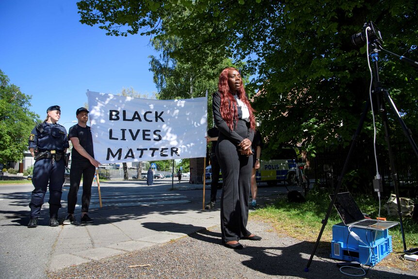 On a bright blue day, you view a woman of African descent speaking into a camera in front of a Black Lives Matter banner.