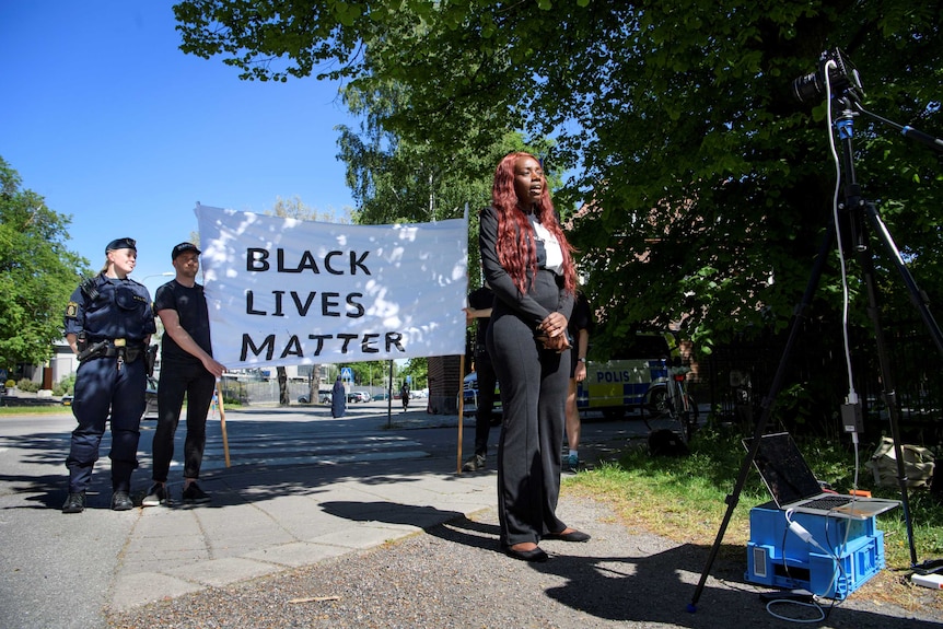 On a bright blue day, you view a woman of African descent speaking into a camera in front of a Black Lives Matter banner.