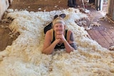 A man lies on a a bed of wool on a shed floor, holding up his head with his hands and smiling at the camera. 