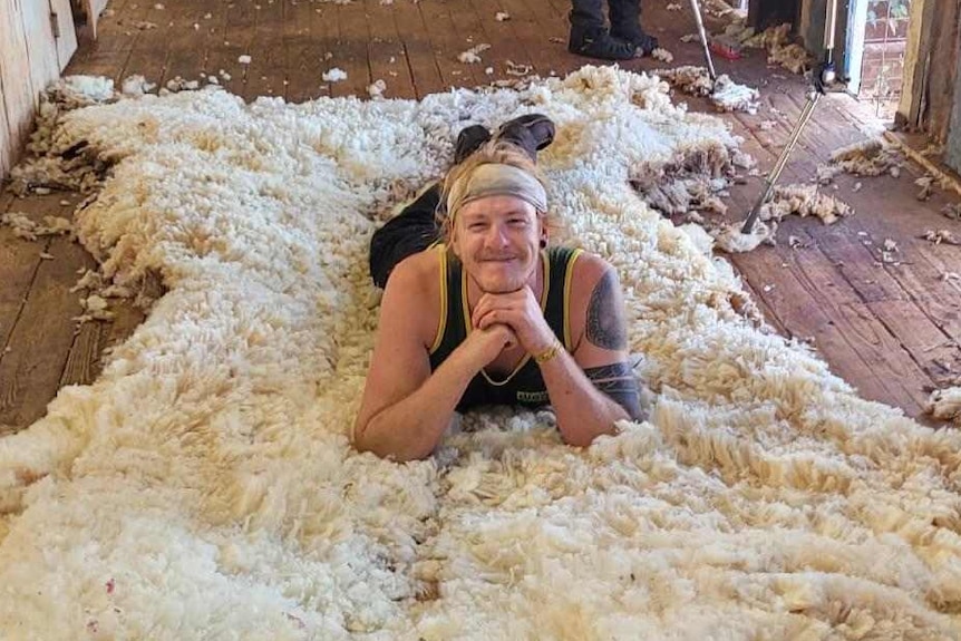 A man lies on a a bed of wool on a shed floor, holding up his head with his hands and smiling at the camera. 