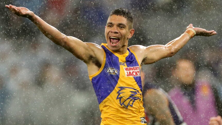 A male AFL player stands with his arms stretched out wide as he smiles in the rain.