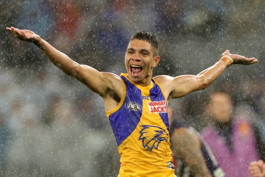 A male AFL player stands with his arms stretched out wide as he smiles in the rain.