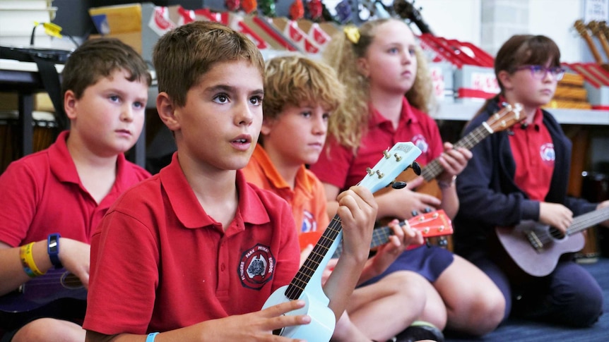 A group of school children sit cross-legged while playing ukuleles.