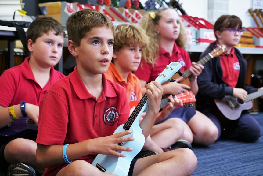 A group of school children sit cross legged while playing ukuleles.