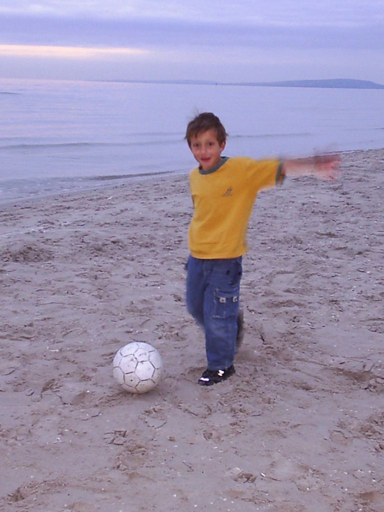 Момче с жълта тениска и сини шорти рита топка в пясъка на плажа