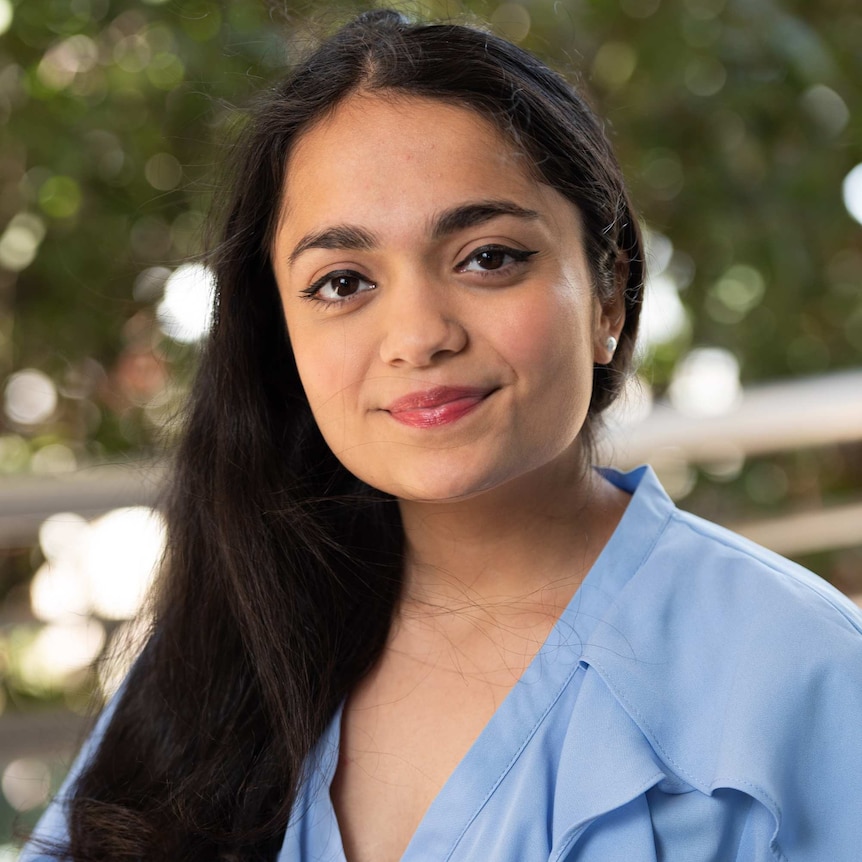 Karishma Luthria looks at the camera smiling in a portrait taken with greenery in the background.