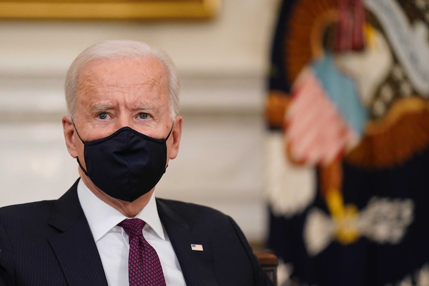An old man in a suit wears a face mask as he looks off camera.