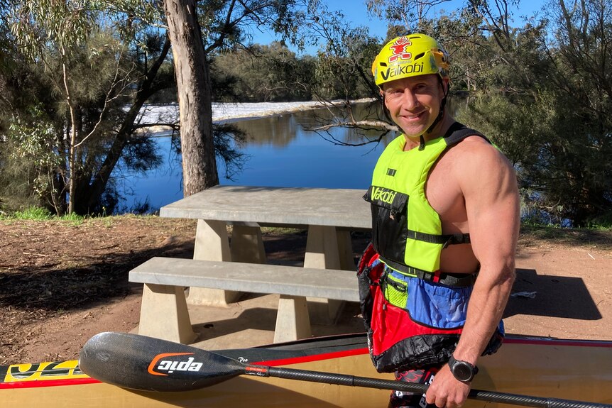 Man wearing life jacket and helmet, holding a kayak standing in front of a river surrounded by trees. 