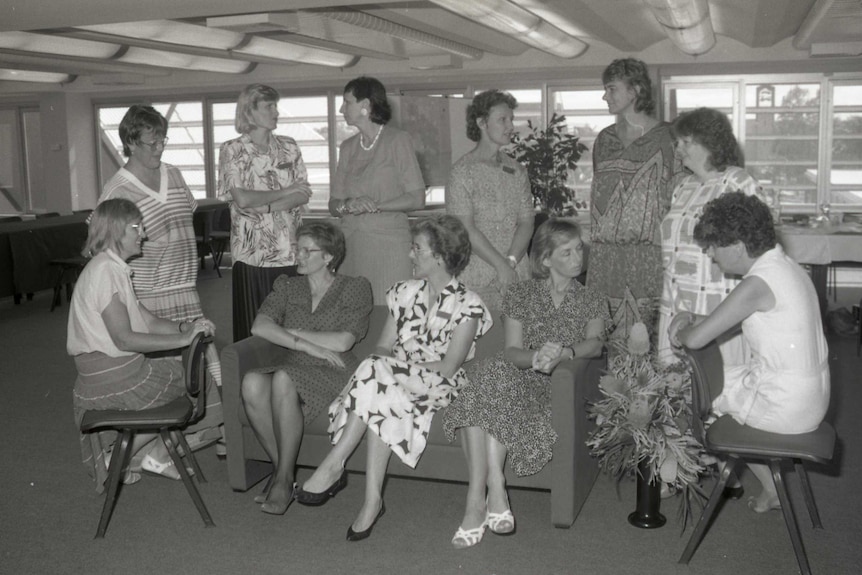 A black and white photo of the women's advisory council; 11 women are pictured sitting or or standing around a chair.