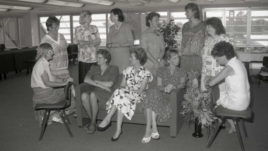 A black and white photo of the women's advisory council; 11 women are pictured sitting or or standing around a chair.