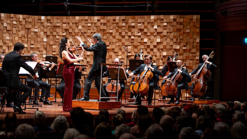 Violinist Ji Won Kim plays violin onstage with an orchestra. She is wearing a glittering long dress.