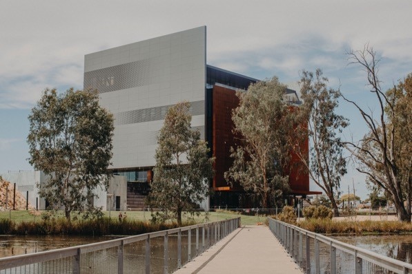 A walkway on a lake with a large grey and rust building in the distance surrounded by trees