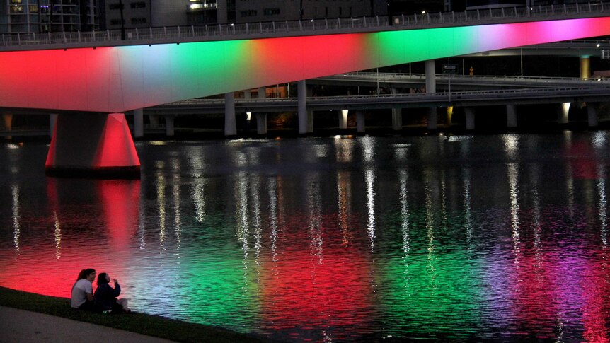 Three people sit on the bank of the Brisbane River, with the Victoria Bridge lit up with various colours.