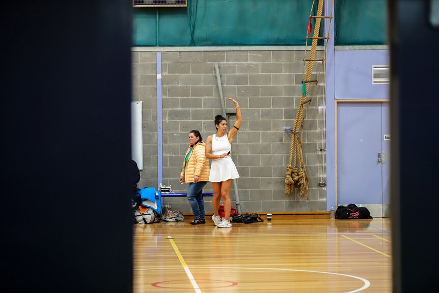 Woman wearing white netball referee outfit gestures on basketball court with a woman walking past behind