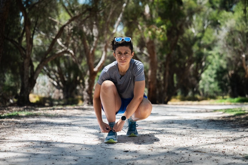 A woman with a determined look crouched down tying her shoe lace on a bushy outdoor running track.