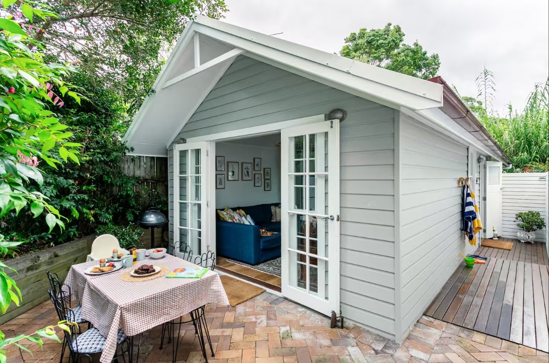 A cute weatherboard grey cottage with french doors opened to a patio with chairs.