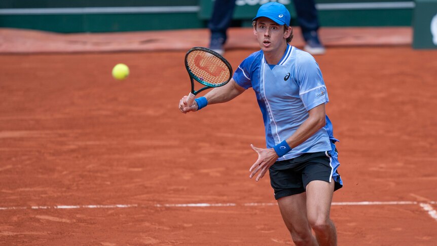 Alex de Minaur, in blue shirt and cap, prepares to hit a forehand on a clay court
