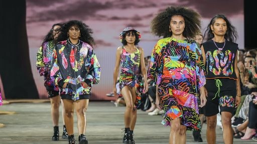 Five people walking down a runway in front of an audience. The models are wearing colourful designs with a black base