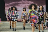 Five people walking down a runway in front of an audience. The models are wearing colourful designs with a black base