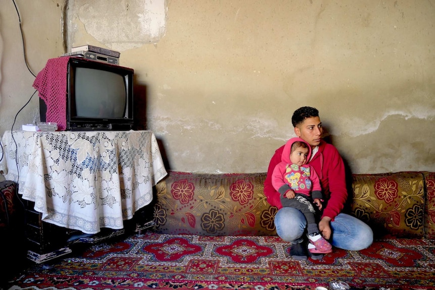 Abood crouches in sparse living room with his daugher