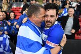 A Greek rugby league player tears up as he stands with his arm around a man wearing a Greek flag.