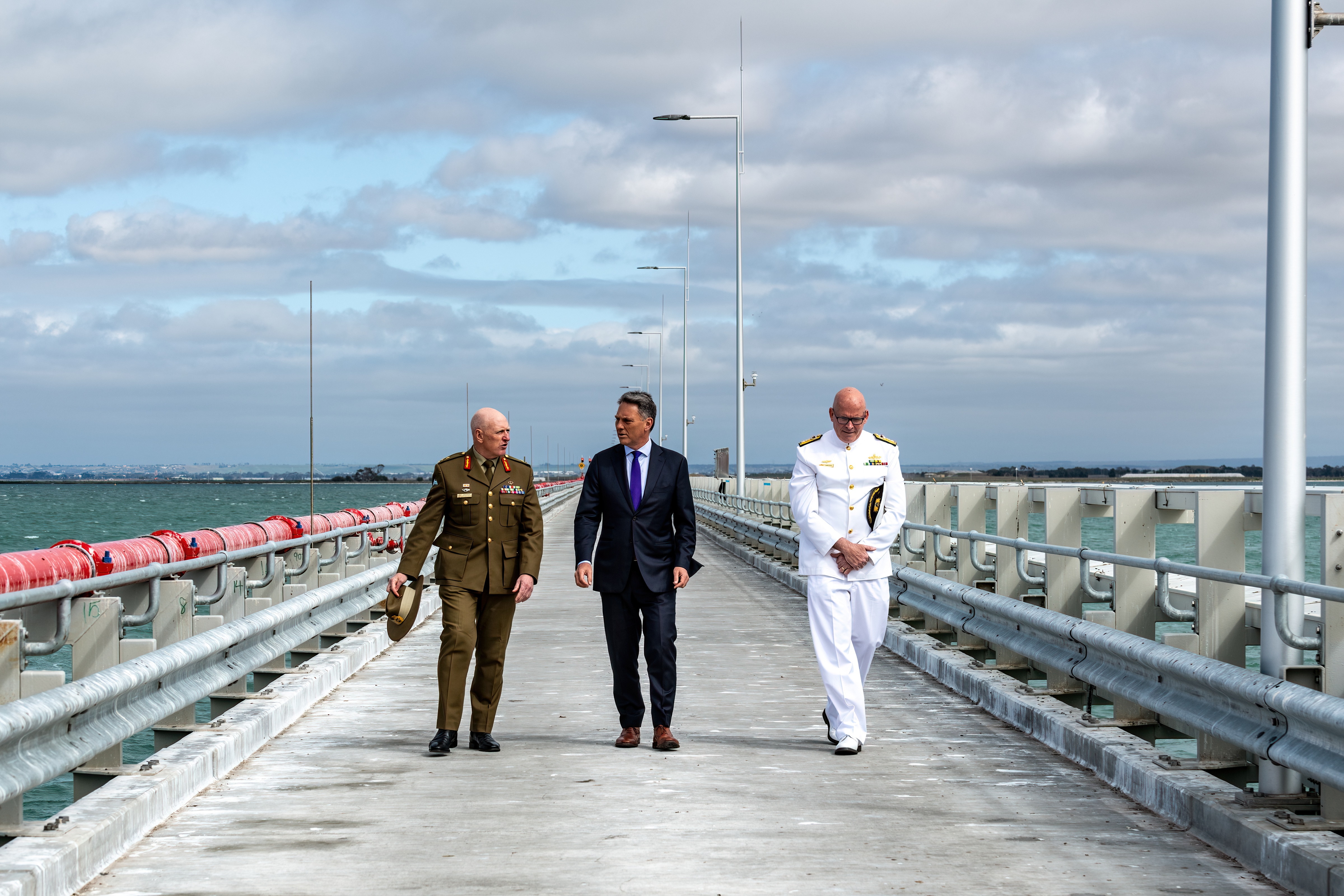 A man in a suit walks along a bare military pier, flanked by two men in military uniform, before a cloudy sky and choppy sea