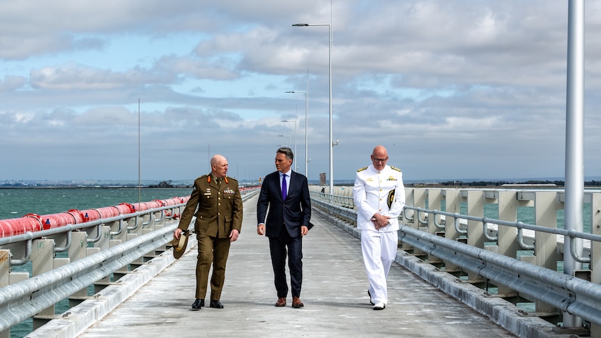 A man in a suit walks along a bare military pier, flanked by two men in military uniform, before a cloudy sky and choppy sea