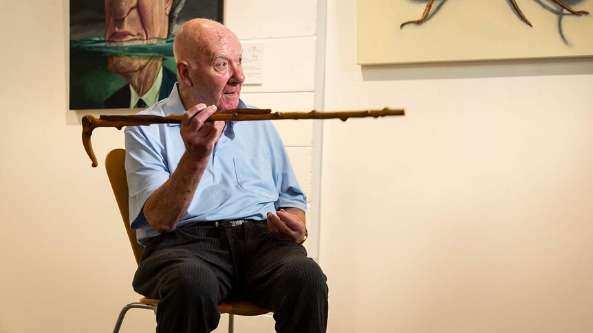 A man in his 80s sits in a chair, pointing his cane, two paintings on the wall behind him.