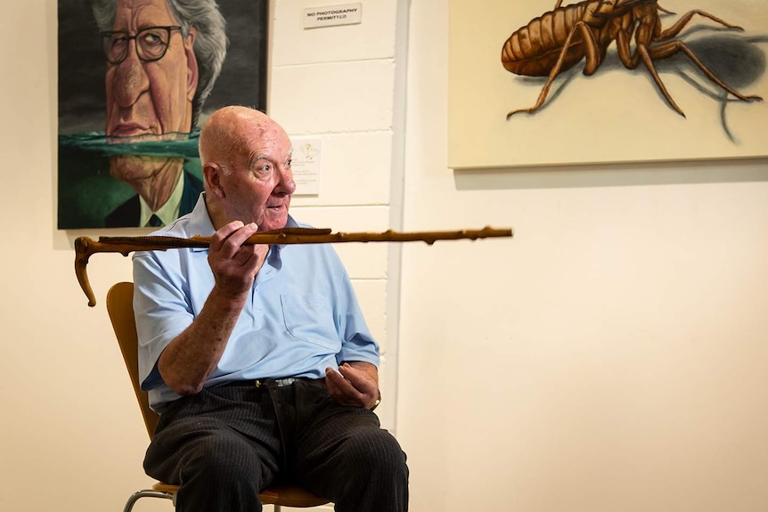 A man in his 80s sits in a chair, pointing his cane, two paintings on the wall behind him.