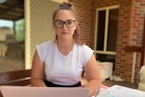 A young woman with glasses sits at an outdoor table while typing on a laptop.