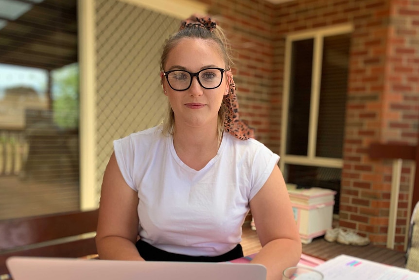 A young woman with glasses sits at an outdoor table while typing on a laptop.